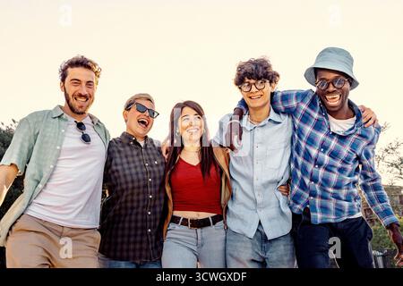 Un groupe joyeux de jeunes amis multiethniques marchant ensemble bras dans bras et riant un jour ensoleillé dans un parc. Banque D'Images
