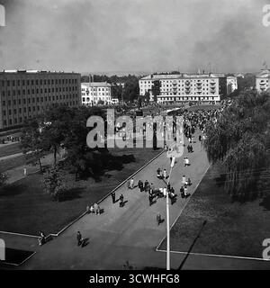 Une vue panoramique à grand angle capture un énorme rassemblement public sur la place de la Révolution d'octobre (maintenant place Soborna) à Sloviansk, en RSS d'Ukraine, lors d'une fête dans toute la ville dans les années 1970 Des foules de gens remplissent la place centrale et les ruelles environnantes, avec le monument de Lénine en son cœur. Flanquée par le bâtiment du conseil municipal et des blocs résidentiels, la scène est un document puissant de la vie communautaire, des rassemblements sociaux et des célébrations d'État dans une ville provinciale soviétique typique pendant l'ère Brejnev Banque D'Images
