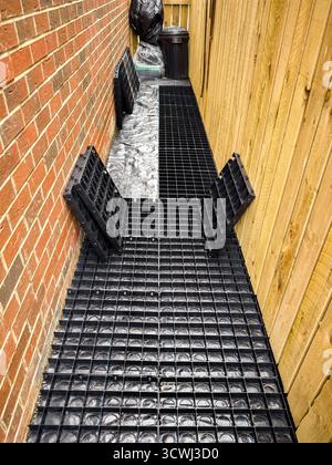 Espace extérieur étroit avec des grilles de drainage noires posées entre le mur de briques et la clôture en bois Banque D'Images