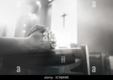 Un croyant priant avec les deux mains ensemble pendant un service religieux, avec la Sainte Bible et la croix de Jésus-Christ en arrière-plan. Banque D'Images