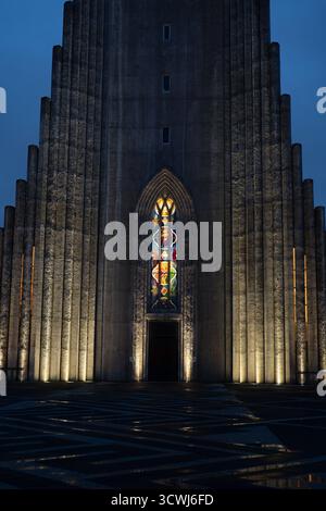 Hallgrimskirkja à Reykjavik, en Islande, présente une façade éclairée avec un grand vitrail, des colonnes géométriques en béton et un premier plan à motifs. Banque D'Images