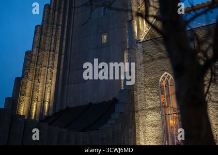 La façade en béton colonnaire de Hallgrimskirkja est éclairée au crépuscule, avec un arbre au premier plan et une lumière chaude d'une fenêtre cintrée visible. Banque D'Images