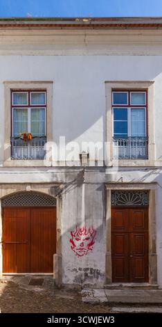 La façade blanche du bâtiment présente deux portes en bois, des fenêtres ornées avec des balcons en fer et un visage graffiti rouge saisissant, le tout sous un ciel bleu clair. Banque D'Images
