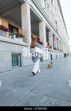 Femme promenant son petit caniche en laisse le long d'Une rue de la ville. Vue arrière, tenue d'été décontractée, ambiance urbaine décontractée. PET Care, Daily routine et Moder Banque D'Images