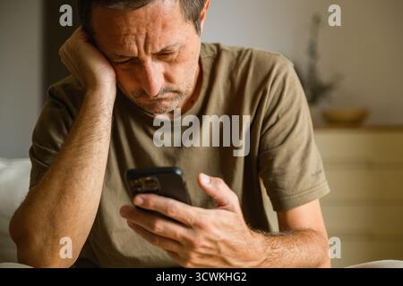 Homme stressé tenant la tête tout en regardant un smartphone, expression frustrée et inquiète à l'intérieur. Mise au point sélective. Banque D'Images