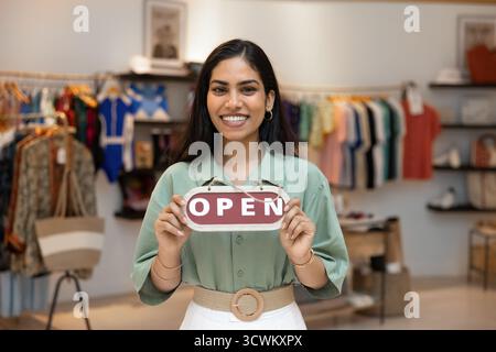 Joyeux et beau jeune directeur de magasin de mode indien montrant la signalétique Open Banque D'Images