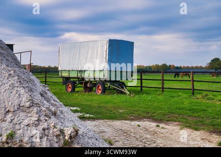 Vintage Green Farm Trailer garé à côté de White Lime pile dans Grassy Field avec clôture en bois et chevaux de pâturage lointains sous Blue Autumn Sky Banque D'Images