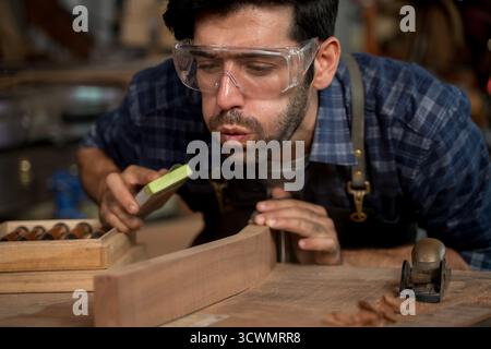Gros plan du charpentier soufflant de la sciure de bois de la surface en bois tout en travaillant dans l'atelier, portant des lunettes de sécurité et un tablier, représentant l'attention à detai Banque D'Images