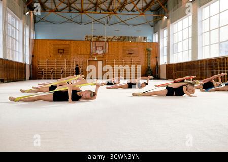 Groupe de filles caucasiennes effectuant des exercices de gymnastique rythmique avec des rubans sur le sol du gymnase, étirant les corps en mouvement synchronisé, axé sur la routine d'entraînement athlétique Banque D'Images