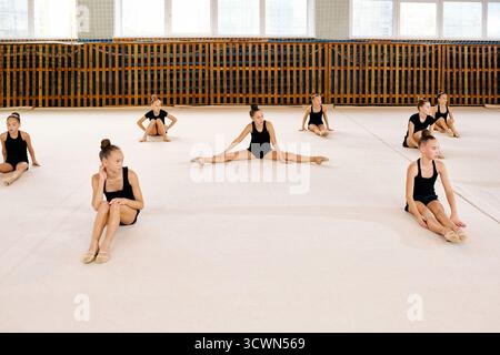 Groupe de jeunes filles caucasiennes préadolescentes assises sur le sol de la salle de gym étirant les jambes pendant la formation de gymnastique rythmique, chaque fille effectuant différents exercices de flexibilité dans une salle de sport spacieuse Banque D'Images