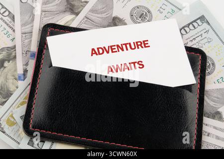 Un portefeuille en cuir noir contenant une carte qui lit AVENTURE ATTEND repose sur une pile de billets de cent dollars américains, signalant un JO planifié Banque D'Images