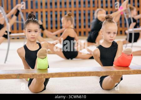 Deux filles caucasiennes effectuant des exercices d'étirement de gymnastique rythmique sur poutre en bois dans le gymnase, portant des justaucorps et des jambières, d'autres filles s'entraînant en arrière-plan, des expressions ciblées Banque D'Images