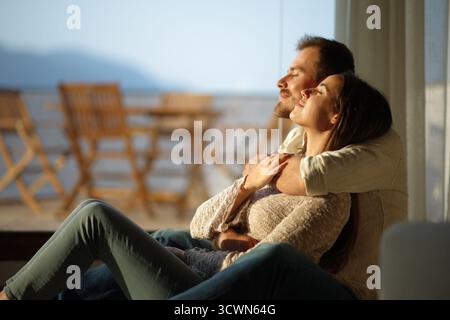 Couple heureux dans l'amour embrassant et se relaxant avec les yeux fermés assis sur le sol à côté d'une fenêtre à la maison Banque D'Images