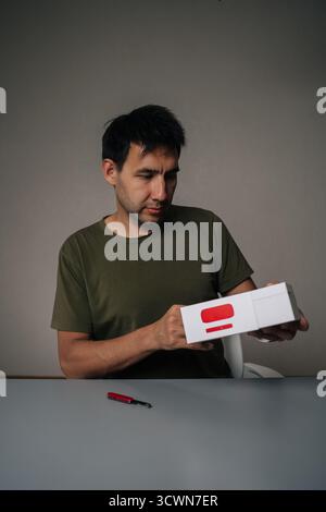 Portrait vertical d'homme blogueur habile déballant la nouvelle cigarette électronique à l'aide d'un petit couteau assis au bureau sur fond gris isolé Banque D'Images