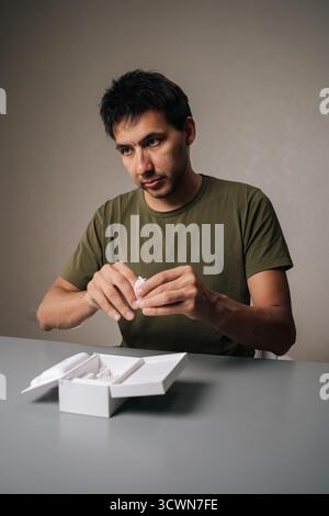 Portrait vertical du client masculin déballant les nouveaux écouteurs sans fil achetés, en retirant de la boîte sur la table grise, exprimant l'excitation Banque D'Images