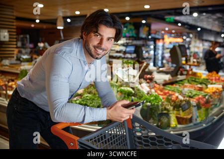 Un homme se penche sur un chariot dans une épicerie, regardant son téléphone Banque D'Images