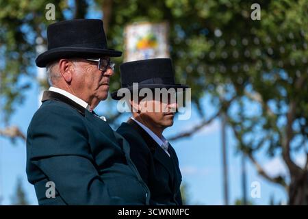 Sevilla, Espagne - 23 avril 23 : deux hommes portant des chapeaux traditionnels et une tenue formelle, assis côte à côte, à l'extérieur avec des arbres en arrière-plan Banque D'Images
