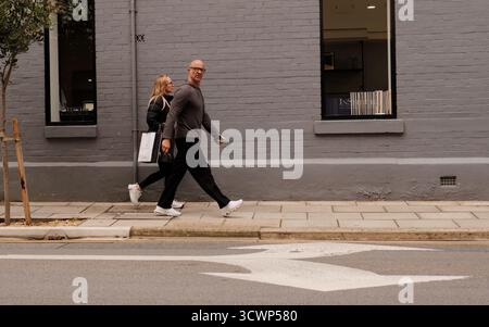 Photo de rue franche, deux adultes marchent sur un trottoir de la ville à côté d'un mur de briques grises. Tons sourds ; un sentiment de vie urbaine et de déplacements quotidiens. Banque D'Images