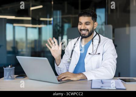 Médecin indien dans un manteau blanc et des gommages menant une consultation médicale en ligne, téléconférence avec un patient, saluant de son lieu de travail de bureau Banque D'Images