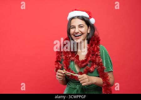 Heureuse femme souriante joyeuse dans la robe verte chapeau de Père Noël avec guirlandes isolées sur fond rouge Banque D'Images