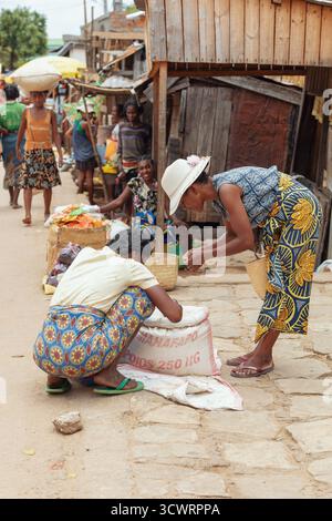 Miandrivazo, Madagascar - 11 octobre 2025 : des femmes malgaches dans lamba colorées échangent des marchandises dans un grand sac sur la rue animée du marché de Miandriva Banque D'Images