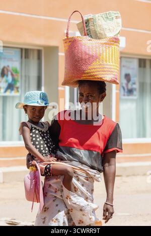 Miandrivazo, Madagascar - 11 octobre 2025 : Portrait d'une mère et d'une fille locales, la femme équilibrant les marchandises sur sa tête dans la ville de Miandrivazo. Banque D'Images