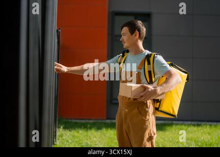 Jeune homme travaillant comme coursier, portant une combinaison marron et un sac de livraison thermique jaune, livrant un petit colis à l'adresse d'un client, tendre la main pour sonner à la porte Banque D'Images