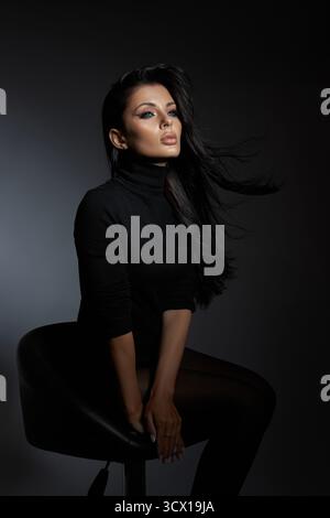 femme confiante s'assoit gracieusement sur un tabouret, ses longs cheveux coulant doucement. un éclairage dramatique souligne ses caractéristiques, créant une atmosphère captivante Banque D'Images