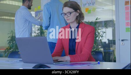 Femme portant un blazer rouge tapant sur un ordinateur portable au bureau, notes adhésives sur un mur de verre, documents imprimés Banque D'Images