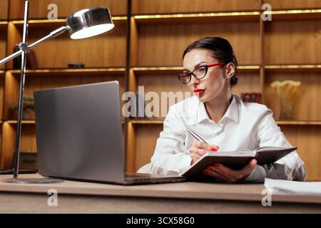 Femme professionnelle prend des notes dans un planificateur tout en utilisant un ordinateur portable, illustrant la planification, la planification, la productivité et le travail à distance dans un bureau moderne. Banque D'Images