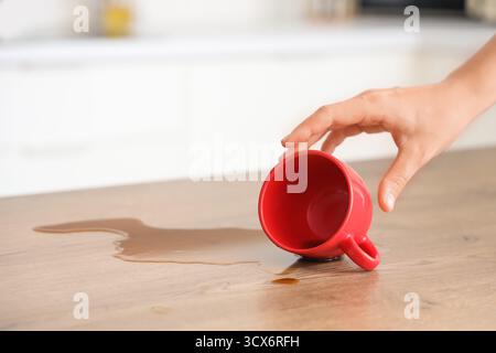 Main femelle renversant le café de la tasse rouge sur la table dans la cuisine, gros plan Banque D'Images