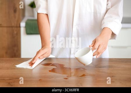 Femme essuyant le café renversé avec une serviette en papier sur la table dans la cuisine, gros plan Banque D'Images