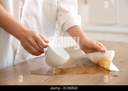 Femme essuyant le café renversé avec une serviette en papier sur la table dans la cuisine, gros plan Banque D'Images