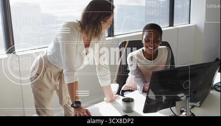 Collaboration deux femmes adultes de taille moyenne portant des chemisiers penchés au-dessus d'un PC au bureau, avec tasse à café Banque D'Images