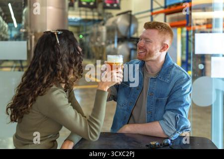 Jeune couple sur une date de brasserie dégustant de la bière artisanale, souriant et détendu dans un taproom industriel moderne, appréciant pintes, conversation et un wee joyeux Banque D'Images