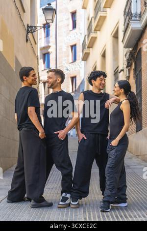 Quatre jeunes amis divers portant des vêtements décontractés noirs interagissent avec joie, sourient et parlent tout en se tenant debout ensemble dans une ruelle urbaine ou St Banque D'Images