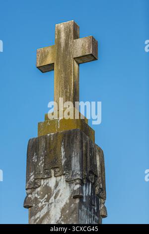 Une croix de pierre altérée au sommet d'un socle se dresse contre un ciel bleu clair. La croix est partiellement recouverte de mousse, indiquant l'âge. Banque D'Images