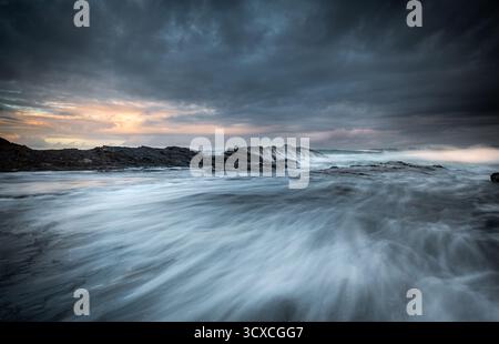 Les vagues se répandent sur une plate-forme rocheuse tandis que les nuages orageux se dressent au-dessus de la mer. L'eau turbulente crée un effet tourbillonnant, semblable à de la brume à travers les roches. Banque D'Images