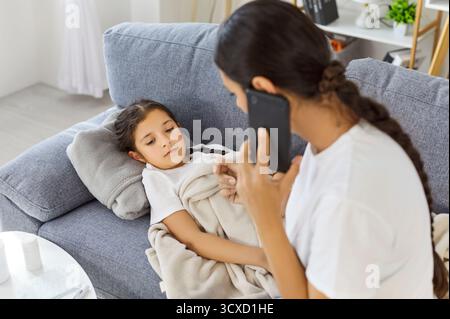 Enfant malade sur canapé avec mère attentionnée appelant le médecin pour l'aide de fièvre Banque D'Images