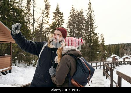 Jeune femme heureuse et homme avec sac à dos en vêtements d'extérieur prenant selfie sur une passerelle en bois dans la neige Banque D'Images