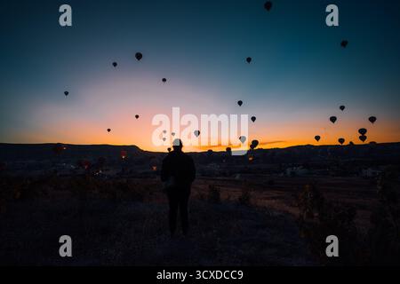 Goreme, Turkiye - 09 octobre 2019 : vue d'une silhouette solitaire sur le ciel ardent de l'aube, ponctuée par les silhouettes flottantes de montgolfières en Cappadoce. Banque D'Images