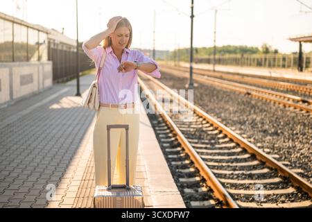 Femme d'affaires mécontente paniquée regardant son horloge tout en se tenant debout avec une valise à la gare. Banque D'Images