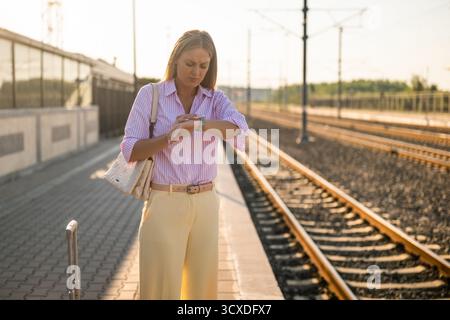 Femme d'affaires mécontente regardant son horloge tout en se tenant debout avec une valise à la gare. Banque D'Images