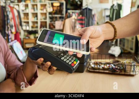 Jeune homme adulte caucasien tenant un smartphone sur le terminal de paiement faisant le paiement sans contact, tandis que femme noire tenant un lecteur de carte dans la friperie avec des vêtements et des accessoires en arrière-plan Banque D'Images