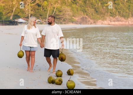 Un couple se promène joyeusement le long d'une plage de sable, se tenant la main et portant des noix de coco. Le soleil brille brillamment, illuminant le littoral paisible et lu Banque D'Images