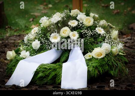 Couronne funéraire traditionnelle avec roses blanches, marguerites et haleine de bébé, couchée sur un sol funéraire fraîchement creusé avec Un ruban blanc blanc de satin Banque D'Images