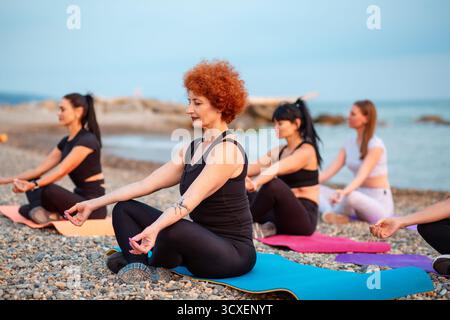 Cours de méditation sur la côte de la mer. Groupe de femmes caucasiennes assis sur des tapis de sport sur la plage de galets. Entraînement de yoga en plein air. Banque D'Images