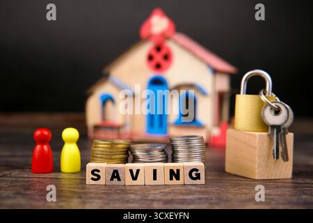 Pièces empilées et blocs de bois orthographiant « SAVING » placés devant un cadenas avec clés et maison miniature, symbolisant l’épargne familiale. Banque D'Images