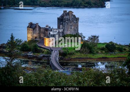 Le château d'Eilean Donan se trouve sur une petite île, reliée par un pont en bois courbe, avec une eau calme et une lumière douce du soir créant un cadre paisible et historique Banque D'Images