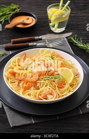 spaghetti aux crevettes dans un bol blanc sur une table en bois sombre avec couverts, sauce, romarin frais et verre d'eau à l'arrière-plan, vue verticale de dessus Banque D'Images
